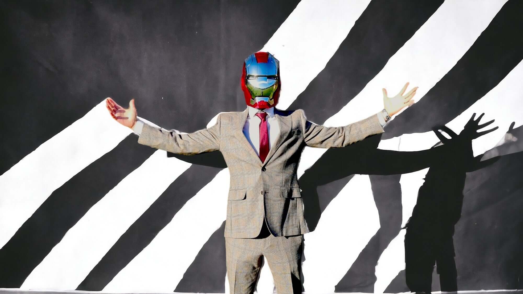 A man in a gray suit, wearing the silver Iron Man MK5 helmet with an artistic black and white background.